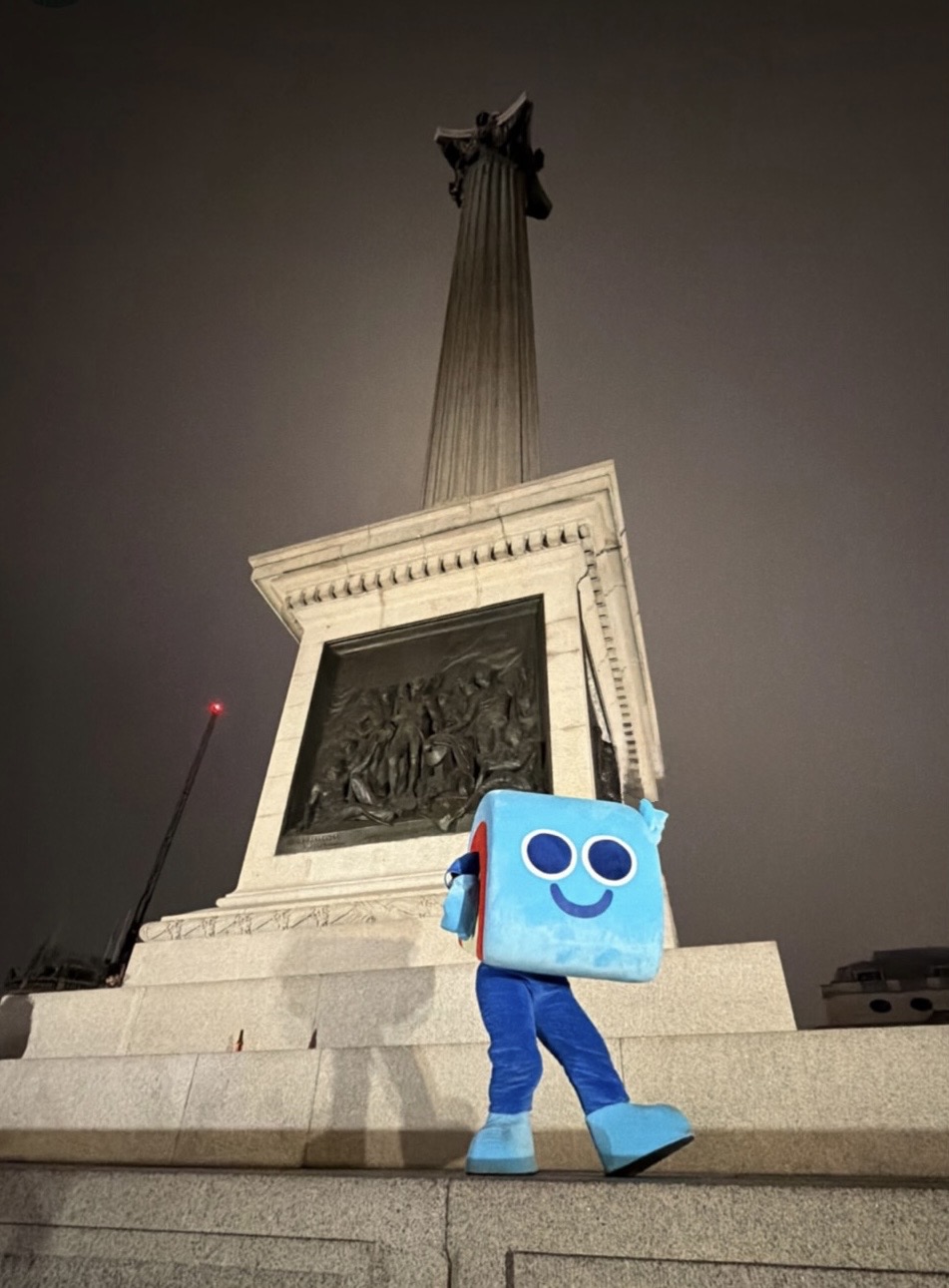Person in a blue Clay cube mascot costume walking the steps of Nelson's Column in Trafalgar Square, London at night